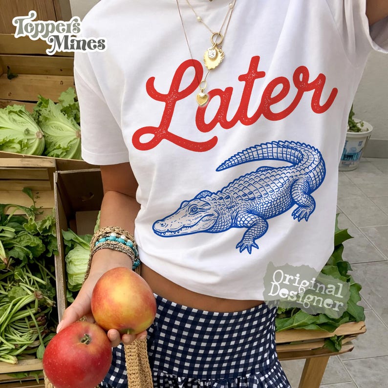 May include: White t-shirt with the word "Later" in red script and a blue alligator illustration. The shirt has a crew neck and short sleeves. The image also shows a person holding two apples.