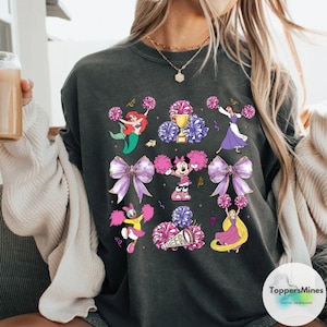May include: A dark grey t-shirt with a graphic print of various Disney characters dressed as cheerleaders. The characters include Ariel, Minnie Mouse, Belle, and Daisy Duck. The graphic also includes purple and pink pom-poms and a cheerleading megaphone.