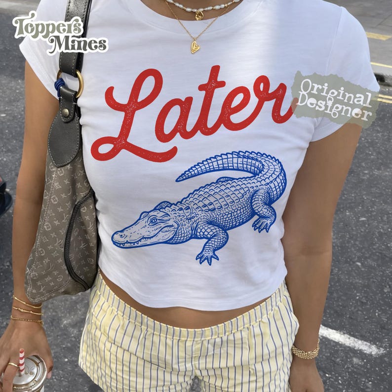 May include: White crop top with the word "Later" in red script and a blue alligator graphic. The shirt has a crew neck and short sleeves. The image also shows a person wearing the shirt with yellow and white striped shorts.