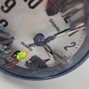 May include: A close-up of a white clock face with black numbers and hands. The clock face is covered in iridescent crystals and a small green figurine. The clock face reads "SHARP" and "QUARTZ".