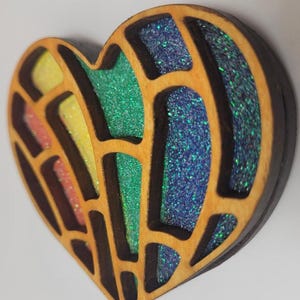 May include: A heart-shaped object featuring a dark brown wooden frame with a cut-out grid pattern. The interior sections are filled with vibrant, multi-colored glitter in shades of yellow, orange, green, and blue, creating a textured appearance.