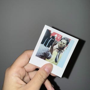 May include: A square instant film photograph featuring a person wearing a textured, light-coloured mask, a yellow apron, and a dark tie. The person is positioned in an indoor setting with a red wall in the background and a person in a striped shirt.