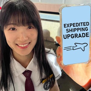 May include: A smartphone with a clear protective case displaying a light blue screen with the black text EXPEDITED SHIPPING UPGRADE and a black line drawing of an aeroplane with motion lines indicating speed, held up for display.