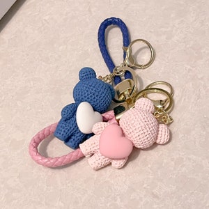 May include: A blue and pink crocheted bear keychain with a heart charm. The keychain is attached to a gold chain and a blue braided cord.