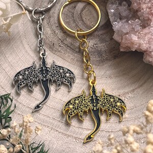 May include: Two keychains with dragon pendants. One keychain has a silver ring and chain, the other has a gold ring and chain. The dragon pendants are black with floral wing designs and the word "LIVE".