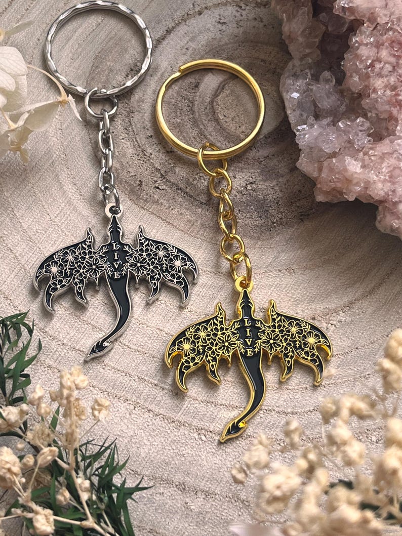 May include: Two keychains with dragon pendants. One keychain has a silver ring and chain, the other has a gold ring and chain. The dragon pendants are black with floral wing designs and the word "LIVE".