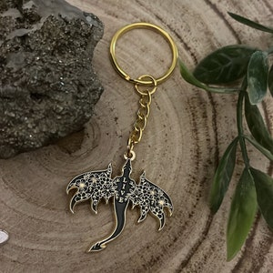 May include: A gold-colored keychain featuring a black dragon pendant with white floral detailing on the wings. The word "LIVE" is vertically displayed on the dragon's body. The keychain is on a gold chain and ring.