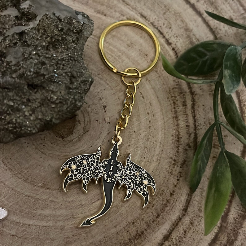 May include: A gold-colored keychain featuring a black dragon pendant with white floral detailing on the wings. The word "LIVE" is vertically displayed on the dragon's body. The keychain is on a gold chain and ring.