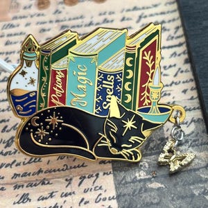 May include: A gold-toned metal enamel pin featuring a black cat sleeping in front of three books labeled Potions, Magic, and Spells. The pin includes a small dangling butterfly charm, a potion bottle, and a candle, all with gold outlines and colorful details.