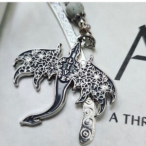 May include: Silver metal bookmark with a dragon design. The dragon has black wings with white floral patterns and the word "LIVE" in white. The bookmark has a gray bead and decorative silver accents.
