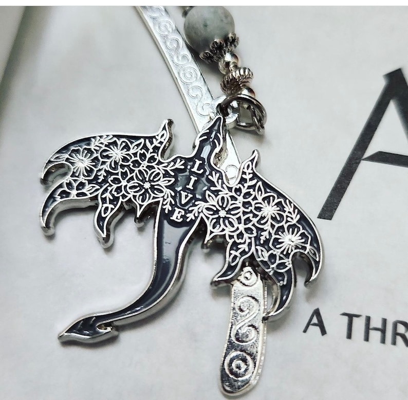 May include: Silver metal bookmark with a dragon design. The dragon has black wings with white floral patterns and the word "LIVE" in white. The bookmark has a gray bead and decorative silver accents.