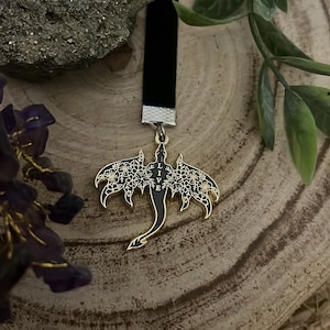May include: A black velvet bookmark with a gold-colored dragon pendant. The dragon has floral wing details and the word "LIVE" vertically down the center. The bookmark is displayed on a wooden surface.