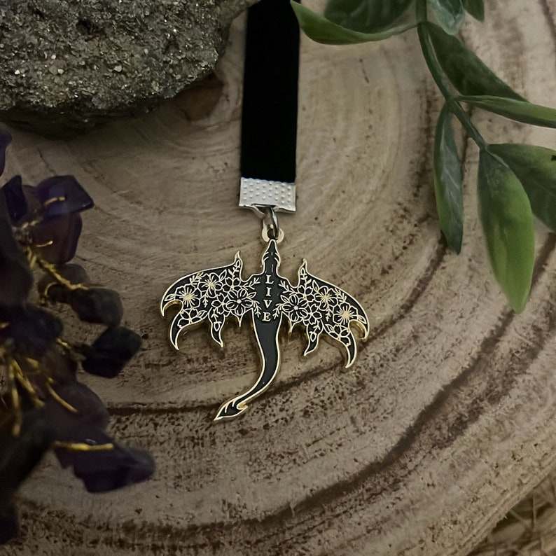May include: A black velvet bookmark with a gold-colored dragon pendant. The dragon has floral wing details and the word "LIVE" vertically down the center. The bookmark is displayed on a wooden surface.