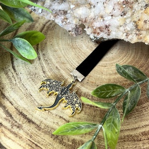 May include: A black velvet choker necklace with a gold and black dragon pendant. The dragon pendant features intricate floral detailing on its wings and body. The necklace is displayed on a wooden surface with green foliage.