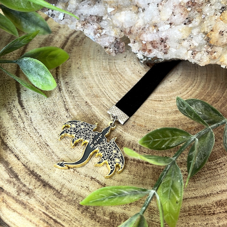 May include: A black velvet choker necklace with a gold and black dragon pendant. The dragon pendant features intricate floral detailing on its wings and body. The necklace is displayed on a wooden surface with green foliage.