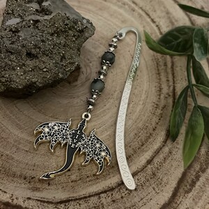 May include: A silver bookmark with a dragon pendant. The dragon is black with white floral details and the word "LIVE". The bookmark has a beaded strand with gray beads and a decorative silver hook.