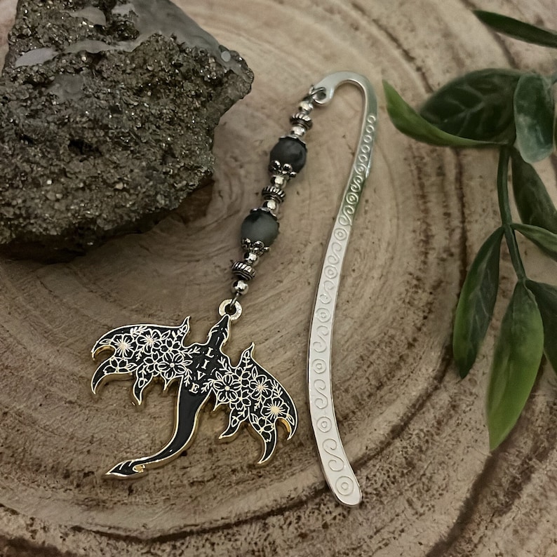 May include: A silver bookmark with a dragon pendant. The dragon is black with white floral details and the word "LIVE". The bookmark has a beaded strand with gray beads and a decorative silver hook.