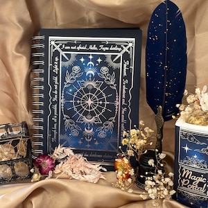 Op de afbeelding: Een zwart spiraalnotitieblok met een zilveren ontwerp met een ster, manen en bloemen. Het notitieblok heeft de tekst "Ik ben niet bang. Hallo, Feyre darling." en "Fireheart. Tot het einde. Je zou kunnen schudden tot de duisternis ons opeist." op de omslag.