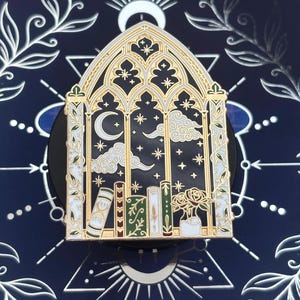 May include: A gold-toned metal enamel pin shaped like a gothic arched window. The design features a dark blue night sky with a crescent moon, stars, and clouds above a row of colorful books and a small vase containing two roses with green leaves.