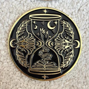 Könnte beinhalten: Schwarzer und goldener Emaille-Pin mit einem Sanduhr-Design und Himmelsmotiven. Der Pin zeigt Monde, Sterne und Hände, mit einem offenen Buch an der Basis. Der äußere Rand ist goldfarben, mit detailliertem Design.