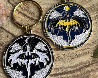 Puede incluir: Dos llaveros de oro con diseños de dragón de esmalte negro y blanco. Los dragones están volando con las alas extendidas. Los llaveros son circulares y tienen un anillo de oro adjunto.