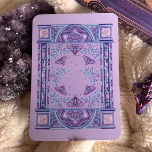 May include: A lavender tarot card with a symmetrical design of moths, leaves, and architectural details in purple and blue hues. The card has rounded corners and a decorative border. The overall theme is mystical and artistic, with a vintage aesthetic.