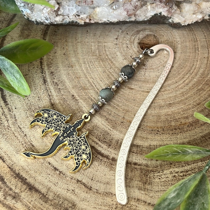 May include: A silver metal bookmark with a dragon charm. The dragon is black with gold accents and floral details. The bookmark has a beaded strand with gray beads. The bookmark is on a wooden surface.