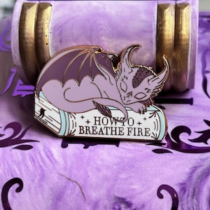 May include: A lavender enamel pin featuring a sleeping dragon with dark purple wings, resting on a book. The book has the text "+ HOW TO + BREATHE FIRE" in white. The pin has gold-colored metal accents.