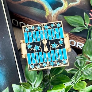 May include: A square gold-toned metal enamel pin featuring two hinged doors that open to reveal a central keyhole. The design includes bright blue vertical panels, black borders, and a pattern of small blue flowers with thin vines.