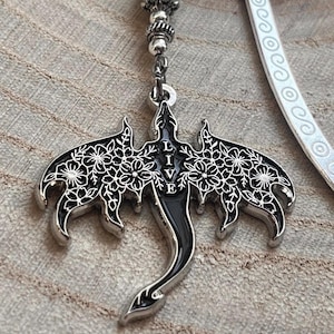 May include: A silver and black dragon pendant with floral wing details and the word "BELIEVE" in the center. The pendant hangs from a beaded chain with a light green stone and a white bookmark.