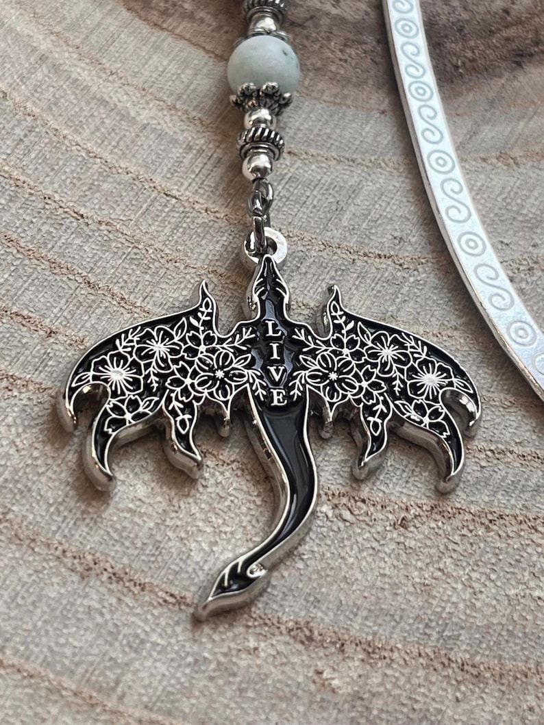 May include: A silver and black dragon pendant with floral wing details and the word "BELIEVE" in the center. The pendant hangs from a beaded chain with a light green stone and a white bookmark.