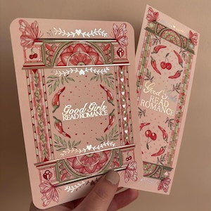 May include: Two pink bookmarks with decorative designs and the text "Good Girls Read Romance." One bookmark features a rectangular design with butterflies, cherries, and chili peppers. The other bookmark has a similar design with a floral border.