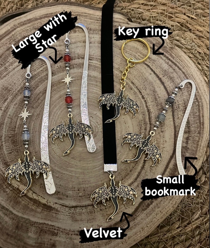 May include: A collection of fantasy-themed accessories, including bookmarks and a key ring. The bookmarks feature ornate metal dragon pendants with the word "LIVE" and decorative beads. One bookmark has a star charm. The key ring has a gold-colored ring.
