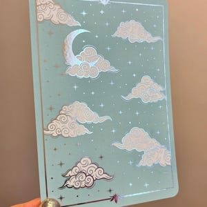 May include: A rectangular card with a light blue background featuring a silver holographic foil design. The illustration depicts a crescent moon, stylised swirling clouds, and small four-pointed stars enclosed within a thin rectangular border.