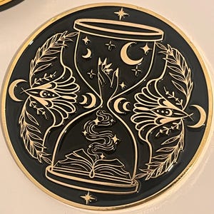 May include: A black and gold circular coaster featuring an hourglass design. The hourglass is filled with stars and moons, and a book is at the bottom. The design is surrounded by leaves and a gold border.