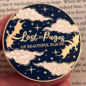 Peut inclure: Épingle ronde en émail bleu marine avec des détails dorés. L'épingle présente le texte "Lost in Pages of Beautiful Places" en lettres dorées, entouré de nuages, d'étoiles et de formes de chauves-souris dorées stylisées.