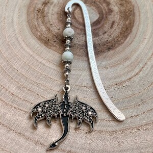May include: A silver metal bookmark with a dragon pendant. The dragon has black enamel and floral cutouts. The pendant hangs from a beaded strand with white and silver beads. The bookmark has a curved top and decorative edge.