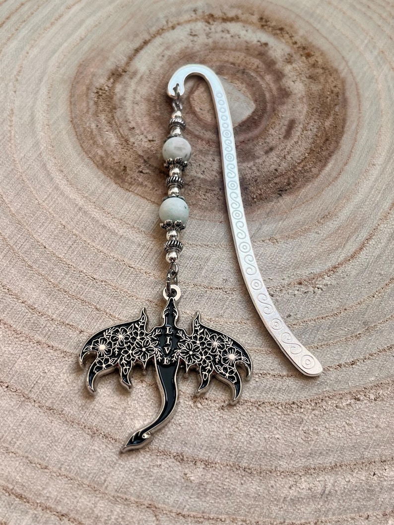 May include: A silver metal bookmark with a dragon pendant. The dragon has black enamel and floral cutouts. The pendant hangs from a beaded strand with white and silver beads. The bookmark has a curved top and decorative edge.