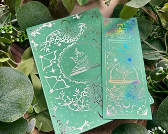 May include: A mint green journal and bookmark set with silver foil accents. The journal features a moth and bow and arrow design, while the bookmark showcases an hourglass and floral elements. The set is surrounded by green foliage.