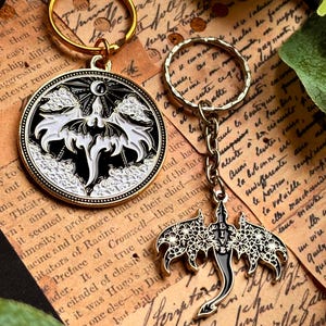May include: Two keychains with gold-colored rings. One is a round pendant with a black and white bat design, the other is a bat-shaped charm with floral details and the word "LIVE". Both are on a vintage paper background.