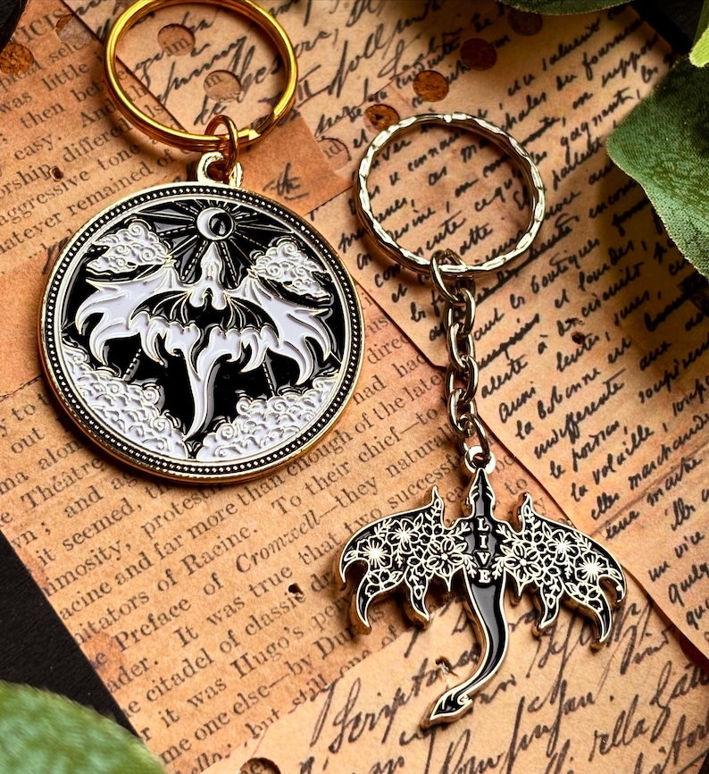 May include: Two keychains with gold-colored rings. One is a round pendant with a black and white bat design, the other is a bat-shaped charm with floral details and the word "LIVE". Both are on a vintage paper background.