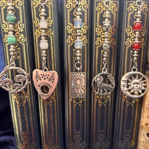 May include: Five decorative metal bookmarks with charms. Charms include a mushroom, a heart with "Witching Hour" text, a tarot card with "The Sun" text, a wolf, and a sun and moon. Each bookmark has a beaded chain with different coloured beads.