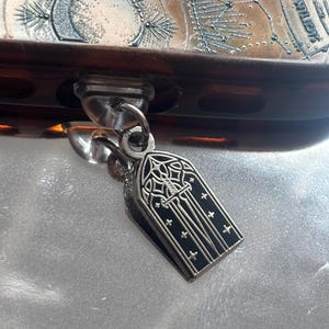 May include: A silver gothic window charm with black enamel and cross details, suspended from a silver ring. The charm is attached to a brown and silver object with celestial map details.