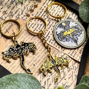 May include: Gold-colored keychains with black and gold dragon designs and a round keychain with a dragon and sun design. One keychain has the text "Fly or Die". The keychains are on a vintage paper background.