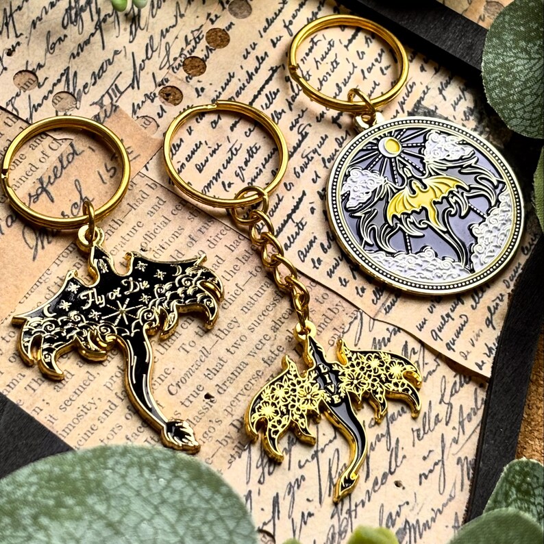 May include: Gold-colored keychains with black and gold dragon designs and a round keychain with a dragon and sun design. One keychain has the text "Fly or Die". The keychains are on a vintage paper background.