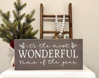 May include: A rectangular, gray wooden sign with white lettering that reads "it's the most WONDERFUL time of the year." The sign is decorated with holly and berries. A small Christmas tree and a decorative ladder are in the background.
