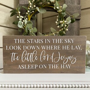 May include: A rectangular wooden sign with the text "THE STARS IN THE SKY LOOK DOWN WHERE HE LAY, the little Lord Jesus ASLEEP ON THE HAY" in white script. The sign is brown and sits on a white surface.