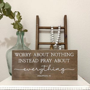 May include: A wooden sign with the text "Worry about nothing instead pray about everything" in white script. The sign is brown and sits on a white surface. A green glass vase and a small wooden ladder are in the background.