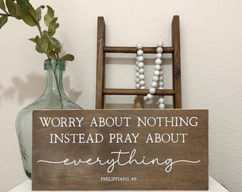 May include: A wooden sign with the text "Worry about nothing instead pray about everything" in white script. The sign is brown and sits on a white surface. A green glass vase and a small wooden ladder are in the background.