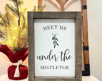 May include: A framed sign with the text "Meet Me Under The Mistletoe" in black script. The sign has a gray wooden frame and a sprig of mistletoe. A small Christmas tree and a decorative wooden window frame are in the background.